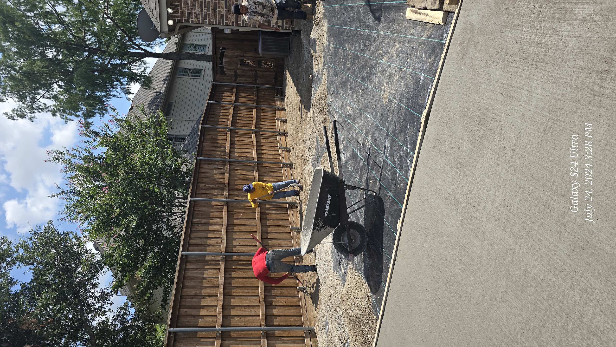 Workers leveling gravel over landscape fabric in a backyard construction project with a wheelbarrow.