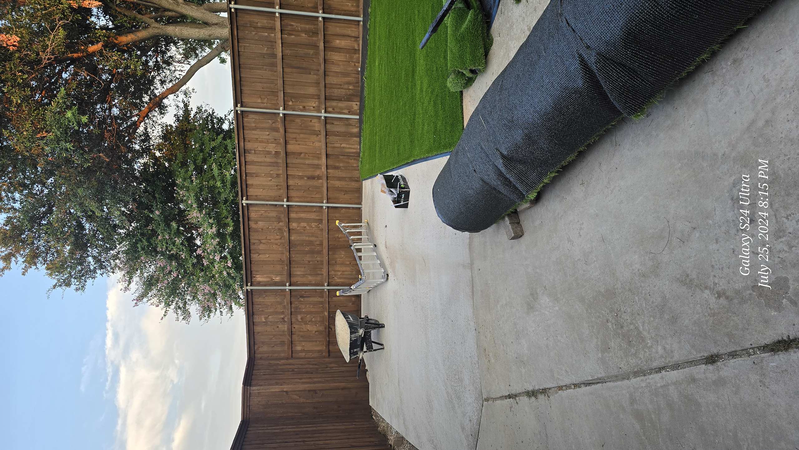 Installation of artificial grass in a backyard with a concrete patio, wheelbarrow, and wooden fence.