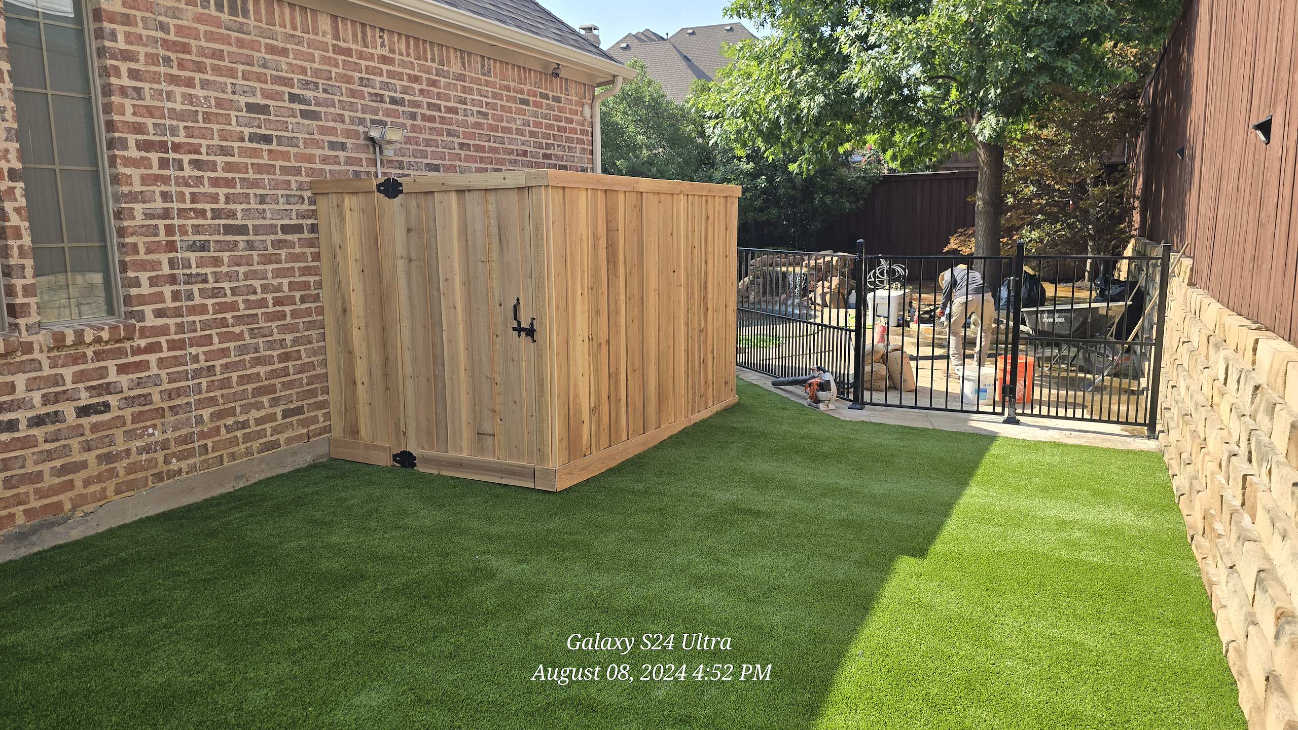 Wooden privacy enclosure on artificial turf beside a brick house and black metal fence.