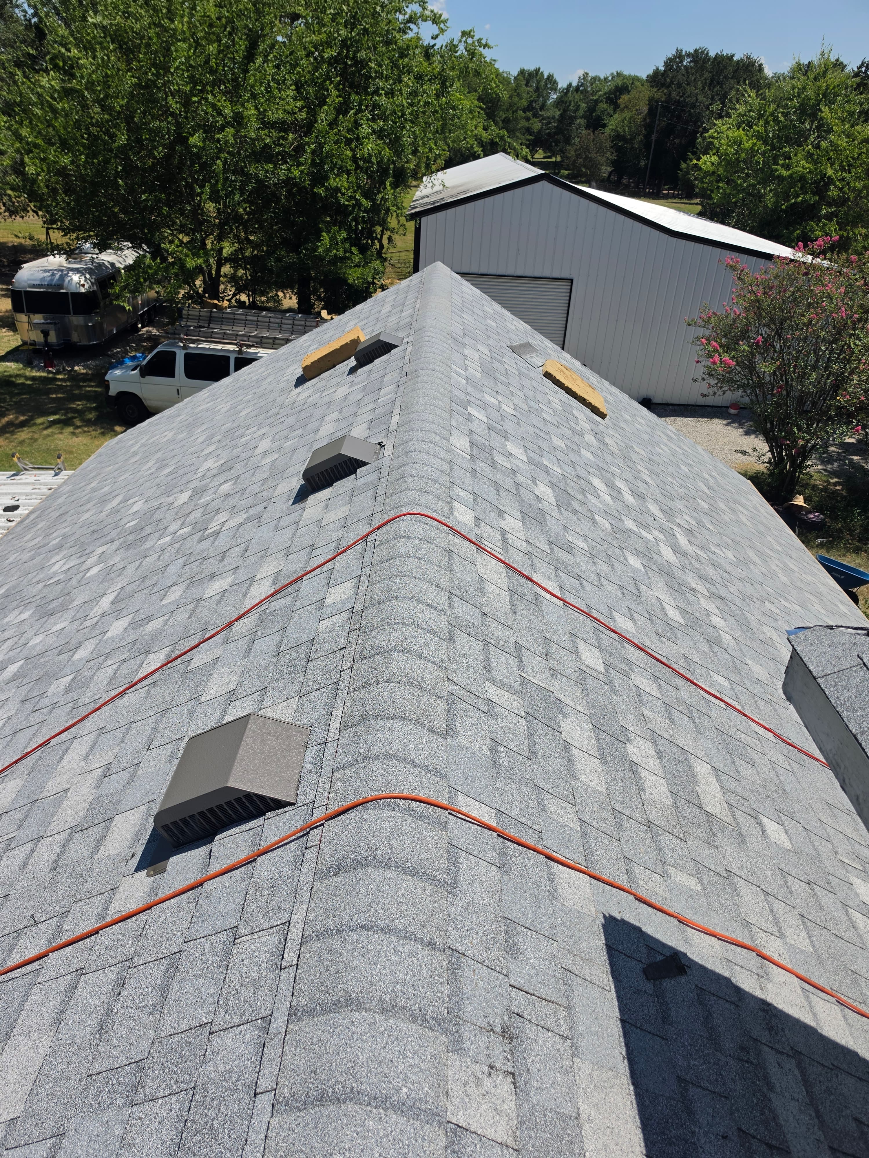 Grey shingled roof ridge with vents and orange cords overlooking a white metal barn.