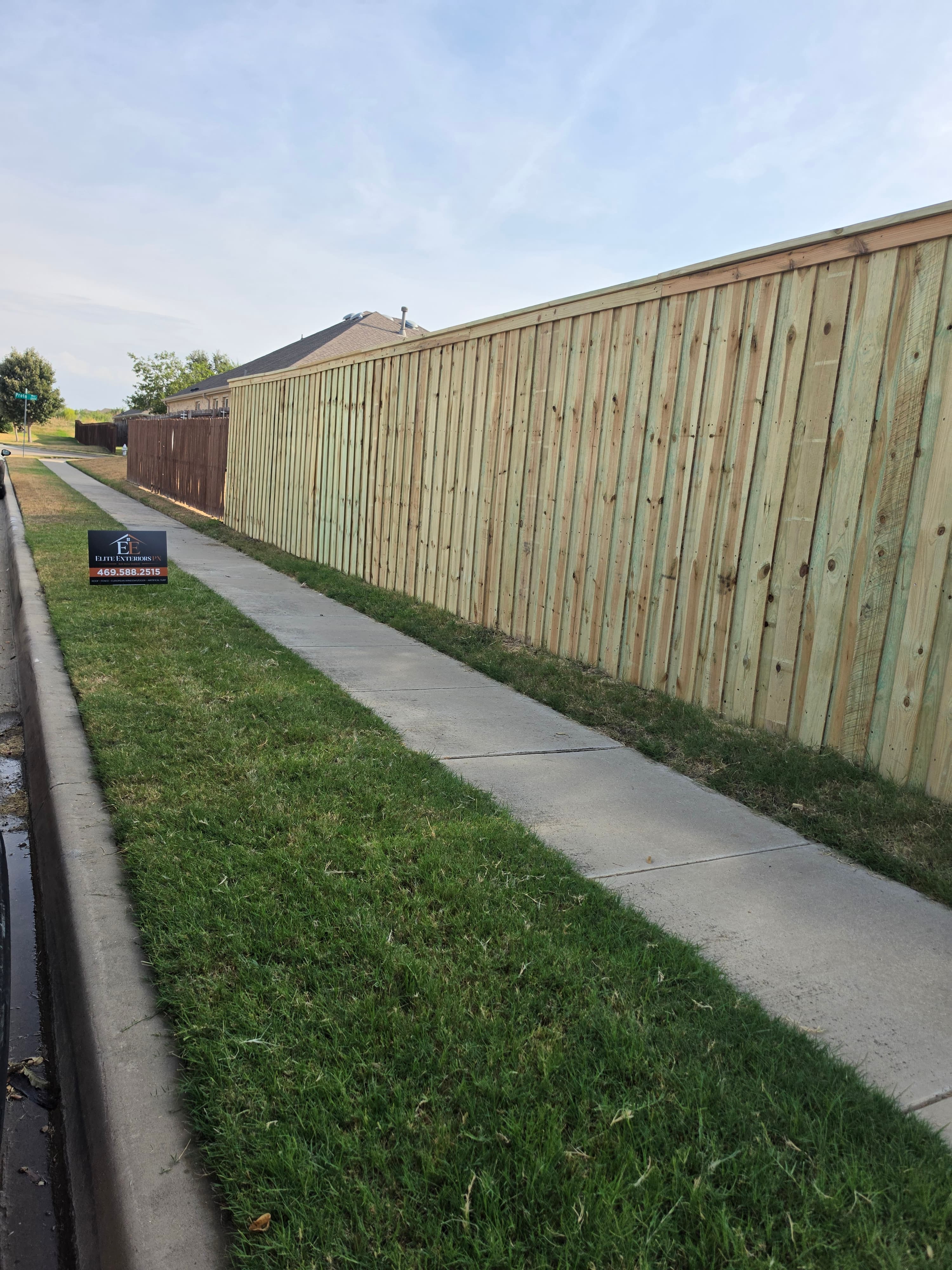 Long wooden fence bordering a concrete sidewalk with an Elite Exteriors sign in the grass.