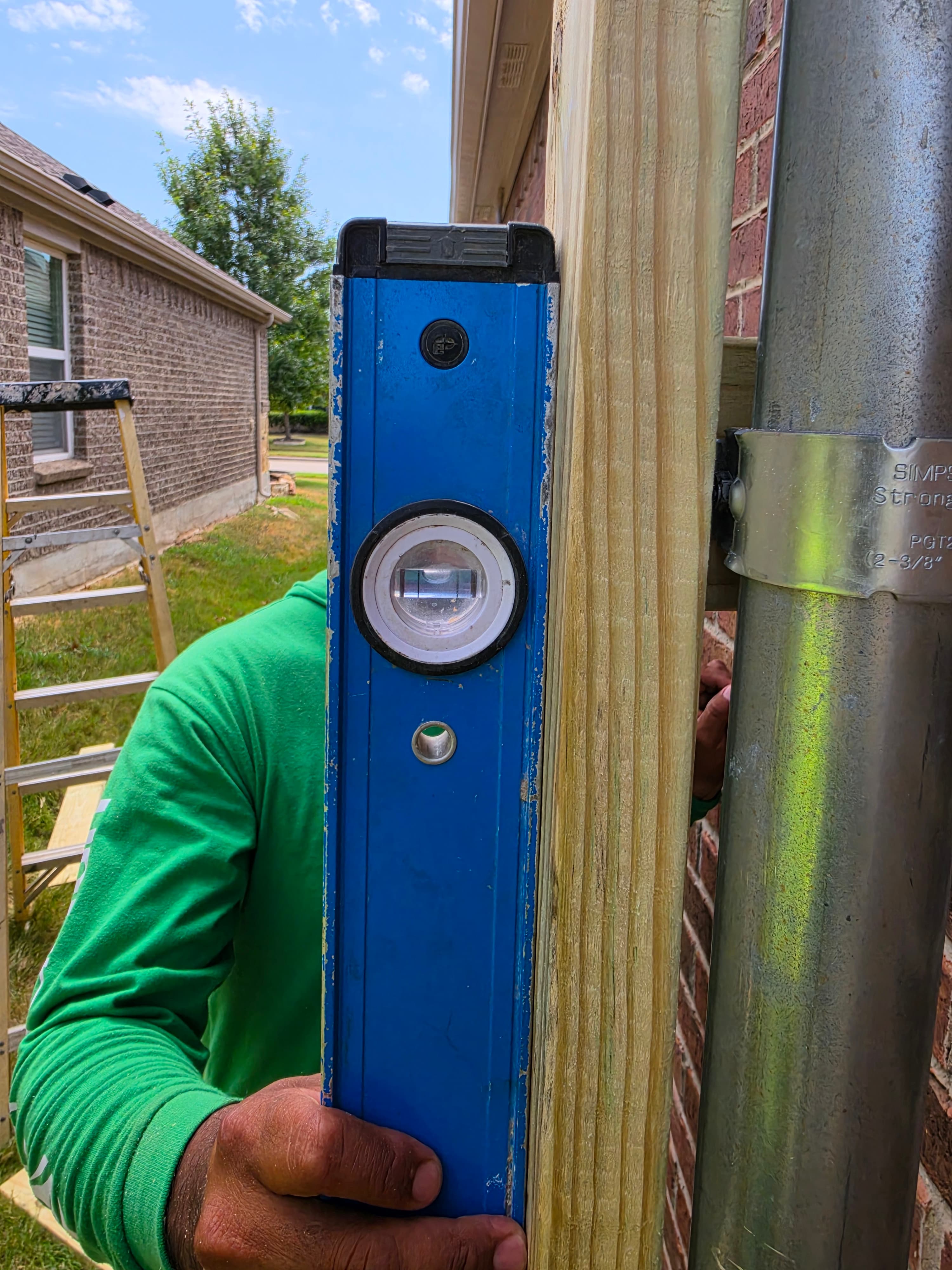 Person in a green shirt holds a blue level against a vertical wooden post.
