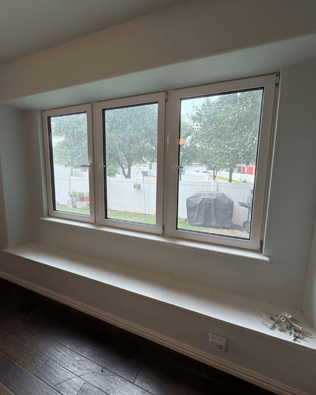 Three-paneled white window with a wide ledge overlooking a backyard with green trees and fence.