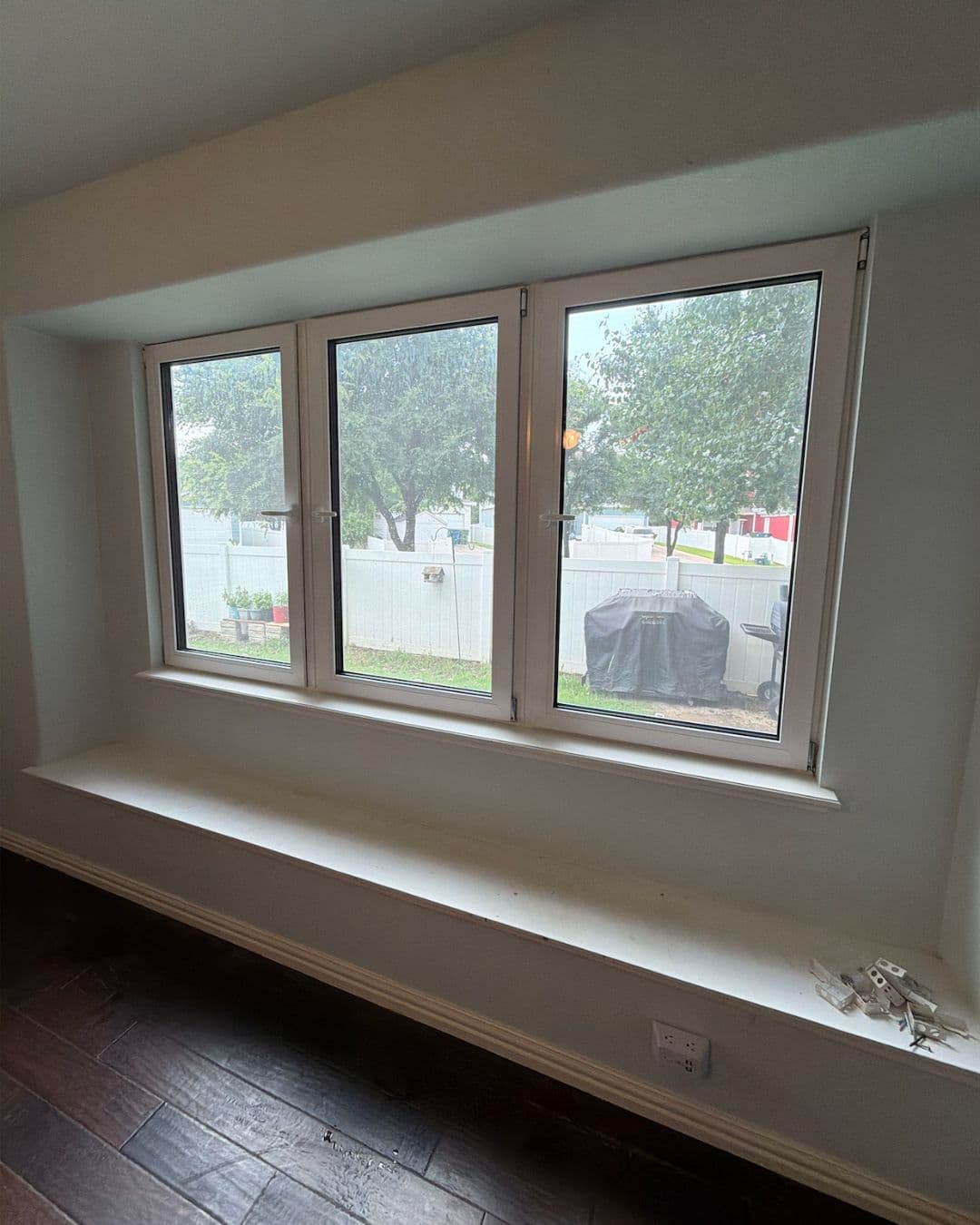Three white windows above a long built-in bench overlooking a fenced backyard with green trees.