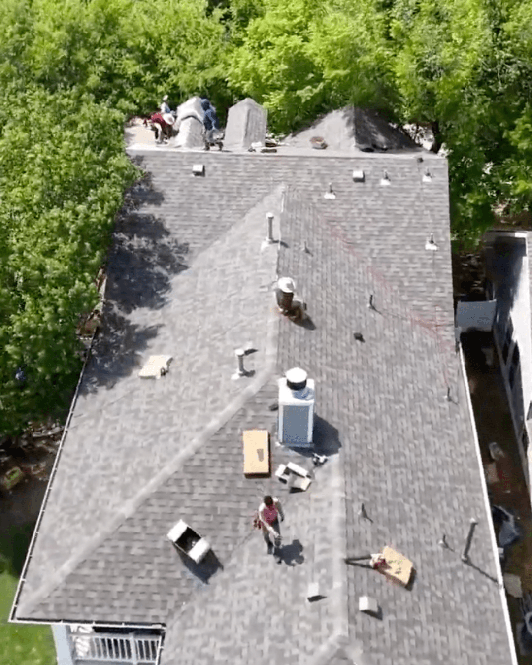 Roofers installing grey shingles on a residential house roof surrounded by lush green trees.