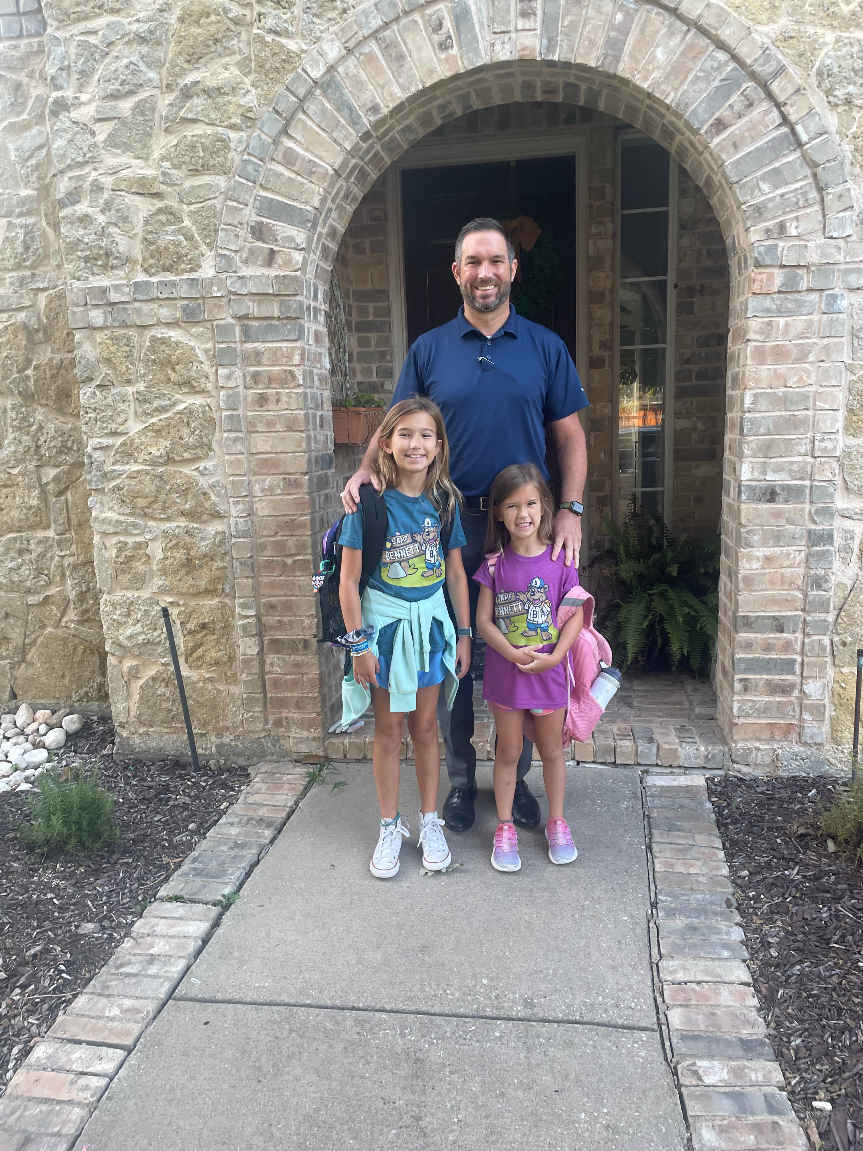 Smiling man and two young girls with backpacks pose before a stone arched entryway.