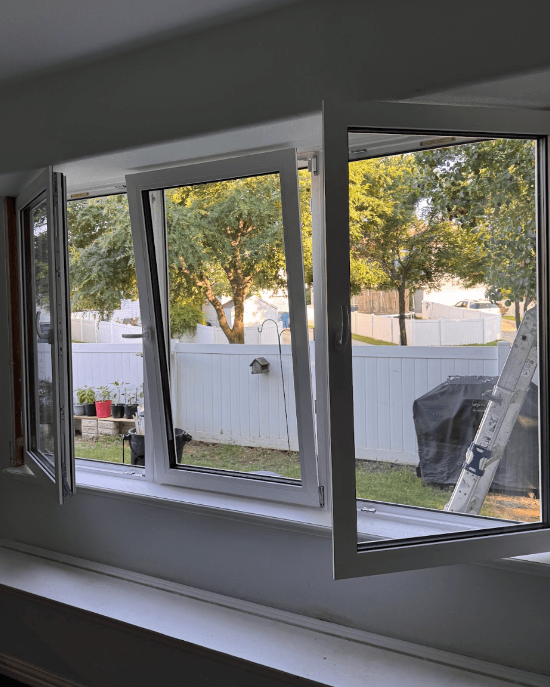 Three white windows open in different positions overlooking a backyard with a white fence.
