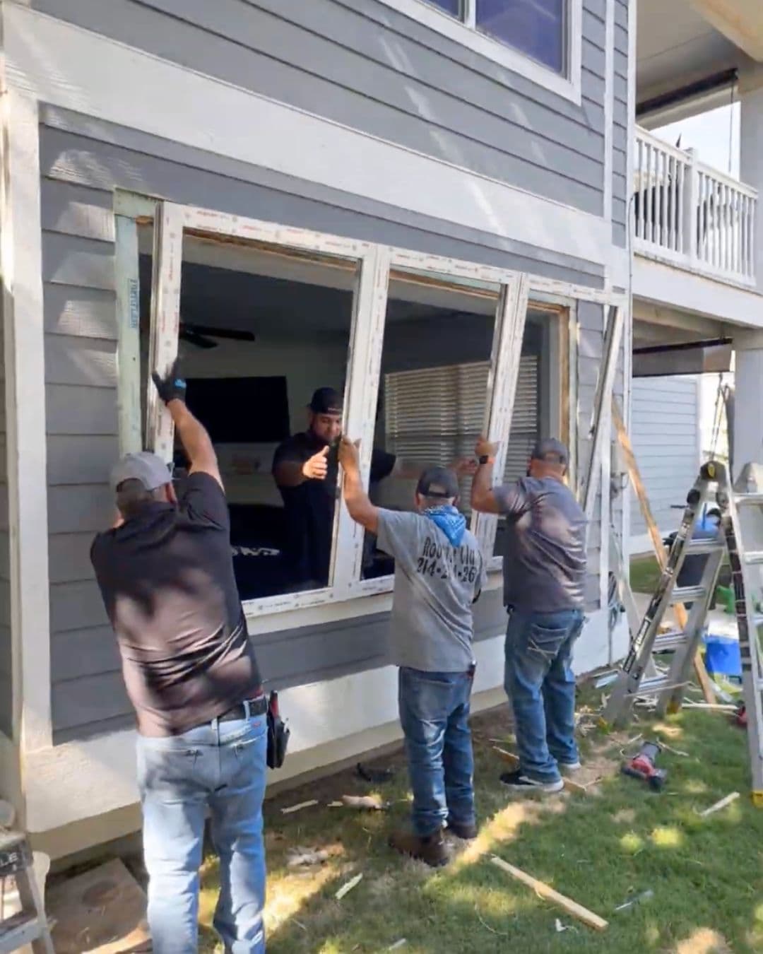 Four workers installing a large white window frame into the gray siding of a house.